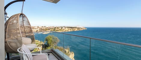 A balcony with a wicker chair, a small table, and a view of the sea and a distant coastline.