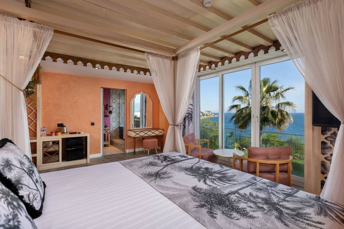 A hotel room with a large bed, a desk with a computer, a chair, a small table, and a balcony with a view of palm trees and the ocean.