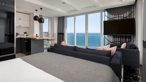 Una habitación de hotel moderna con una cama grande, una zona de estar con vista al mar, un comedor y un televisor montado en la pared.