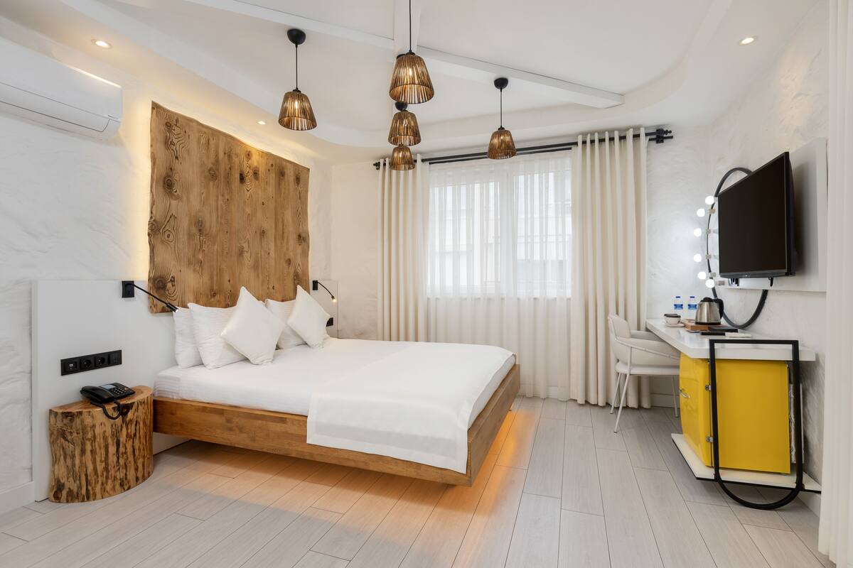 A modern hotel room with a wooden headboard, a bed, a desk with a TV, and a yellow cabinet.