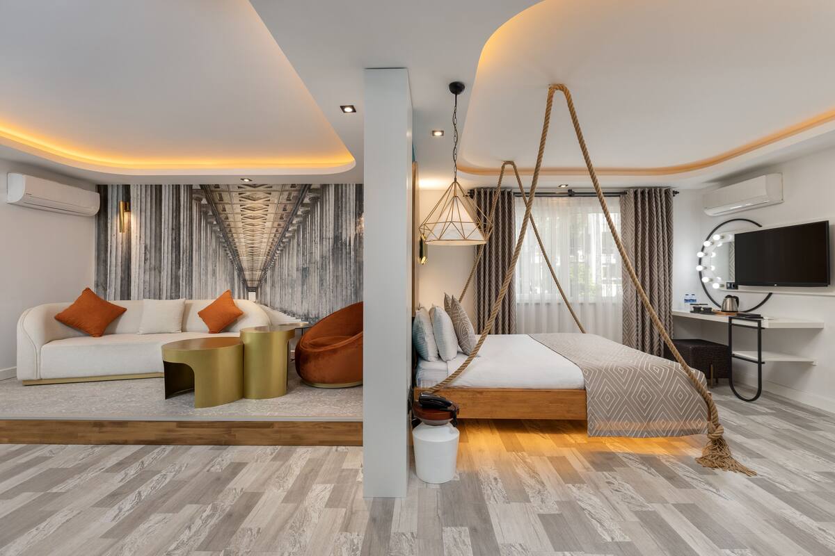 A modern hotel room with a sofa, a bed with a rope swing, a desk with a mirror, and a television.