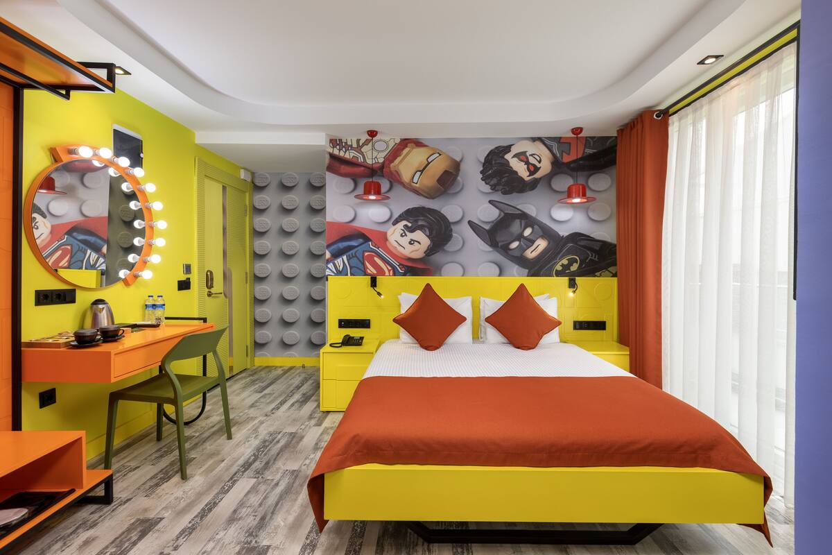 A hotel room with a bed, a desk, a chair, a mirror with lights, and a wall with superhero figures.
