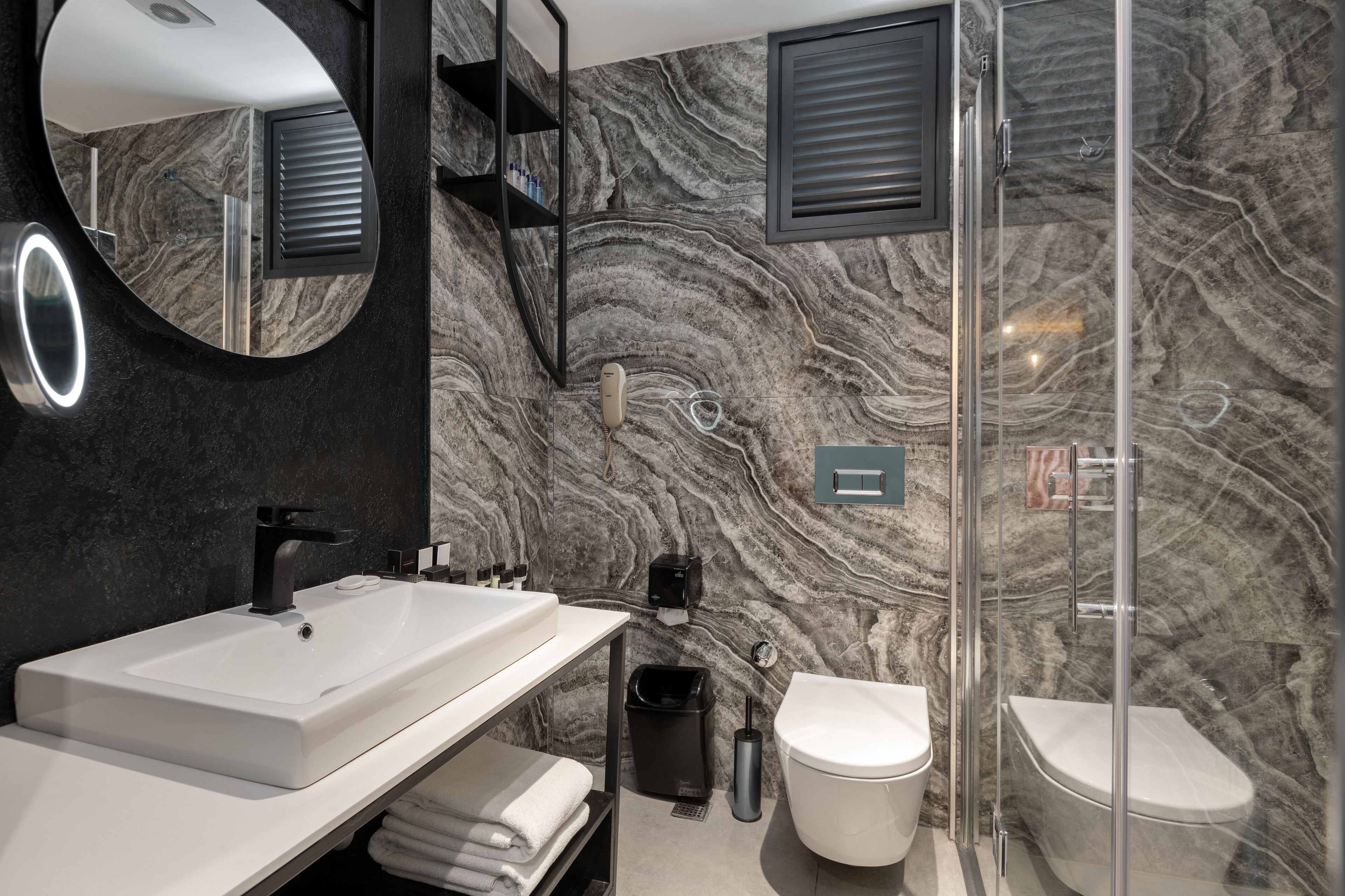 A modern bathroom with a marble backsplash, a white sink, a black faucet, a glass shower enclosure, and a toilet.