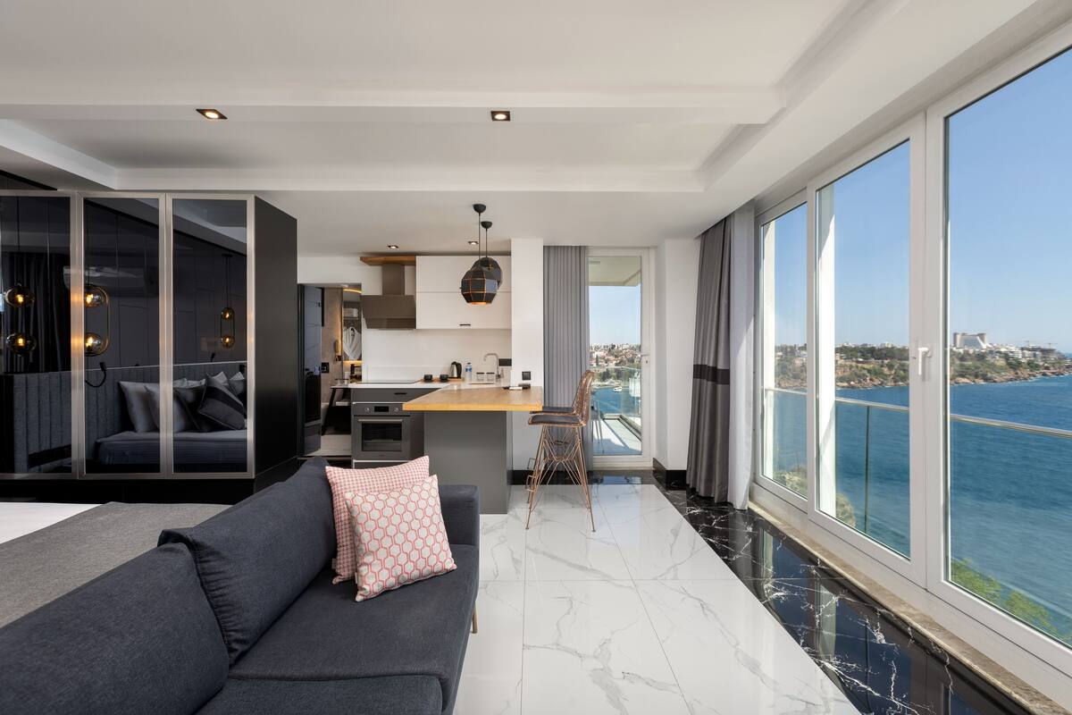 A modern living room with a sofa, a dining area, and a kitchen. There is a large window with a view of the sea.