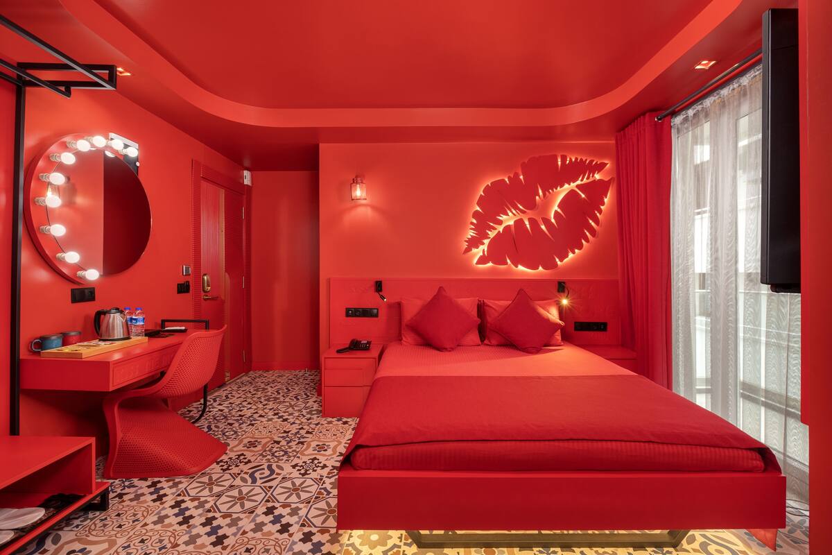 A red-themed bedroom with a large bed, a vanity with a mirror and lights, a patterned floor, and a decorative wall piece.