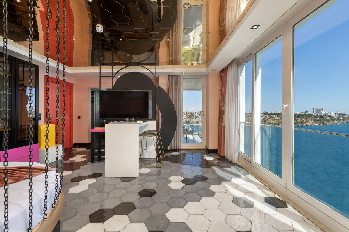 A modern interior with a hexagonal tiled floor, a suspended bed, a TV, and a balcony with a view of the sea.