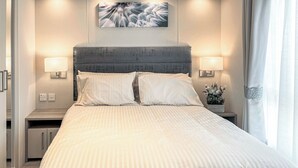 Un lit bien fait, recouvert d’une couvre-lit à rayures, deux lampes de chevet, un tableau abstrait accroché au mur et une table de chevet ornée d’une petite plante.