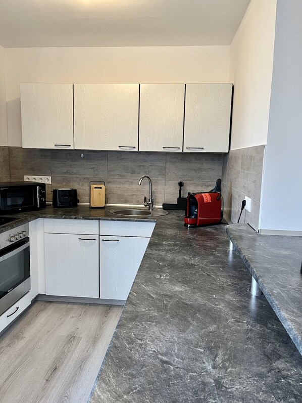 A modern kitchen with white cabinets, a granite countertop, a built-in oven, a microwave, a sink, and a red coffee maker. - Fireball: Luxury Loft | Spa | Workspace | Balcony (Bad Staffelstein)