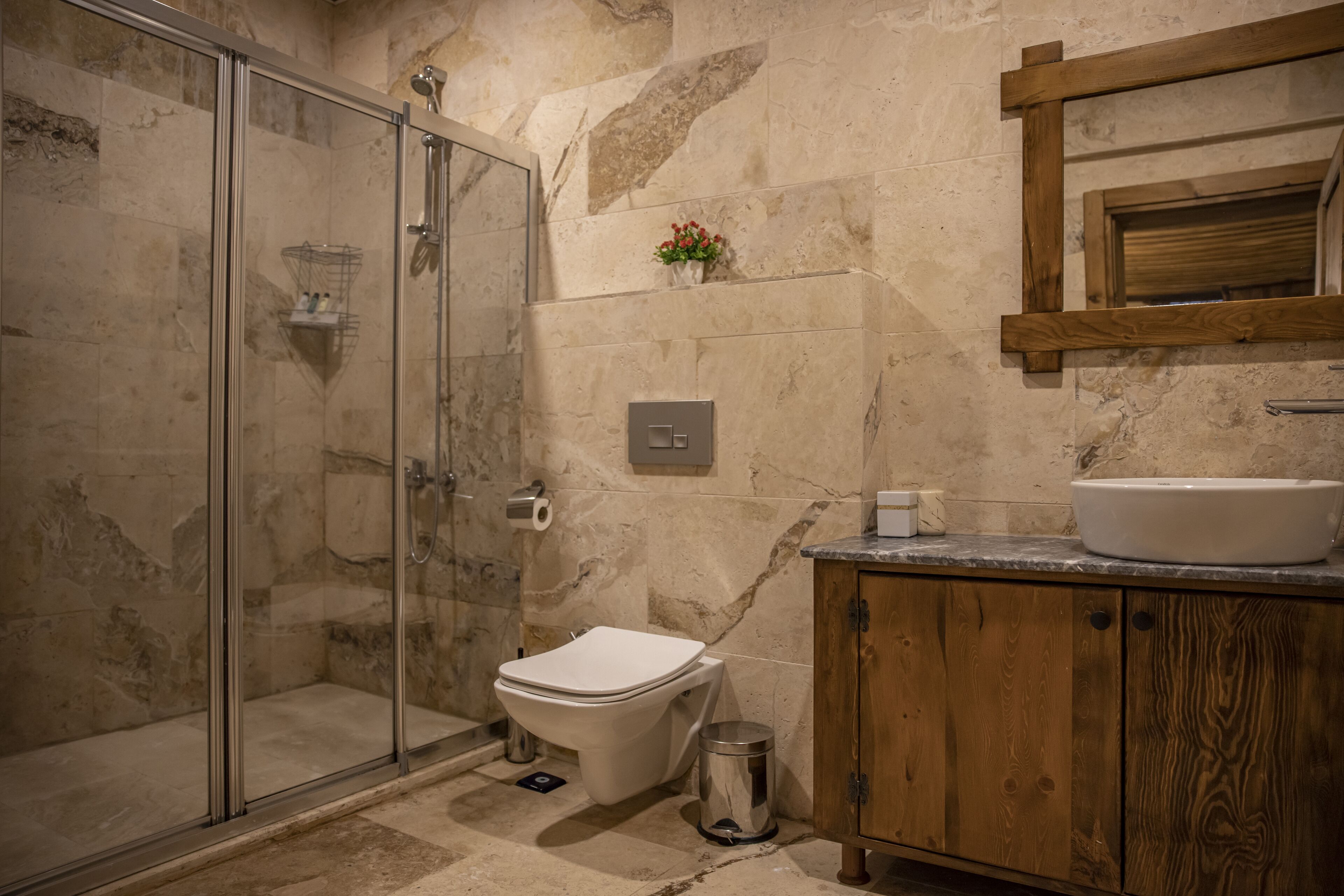 Un baño con ducha de vidrio, inodoro blanco, mueble de madera y pared de mármol.