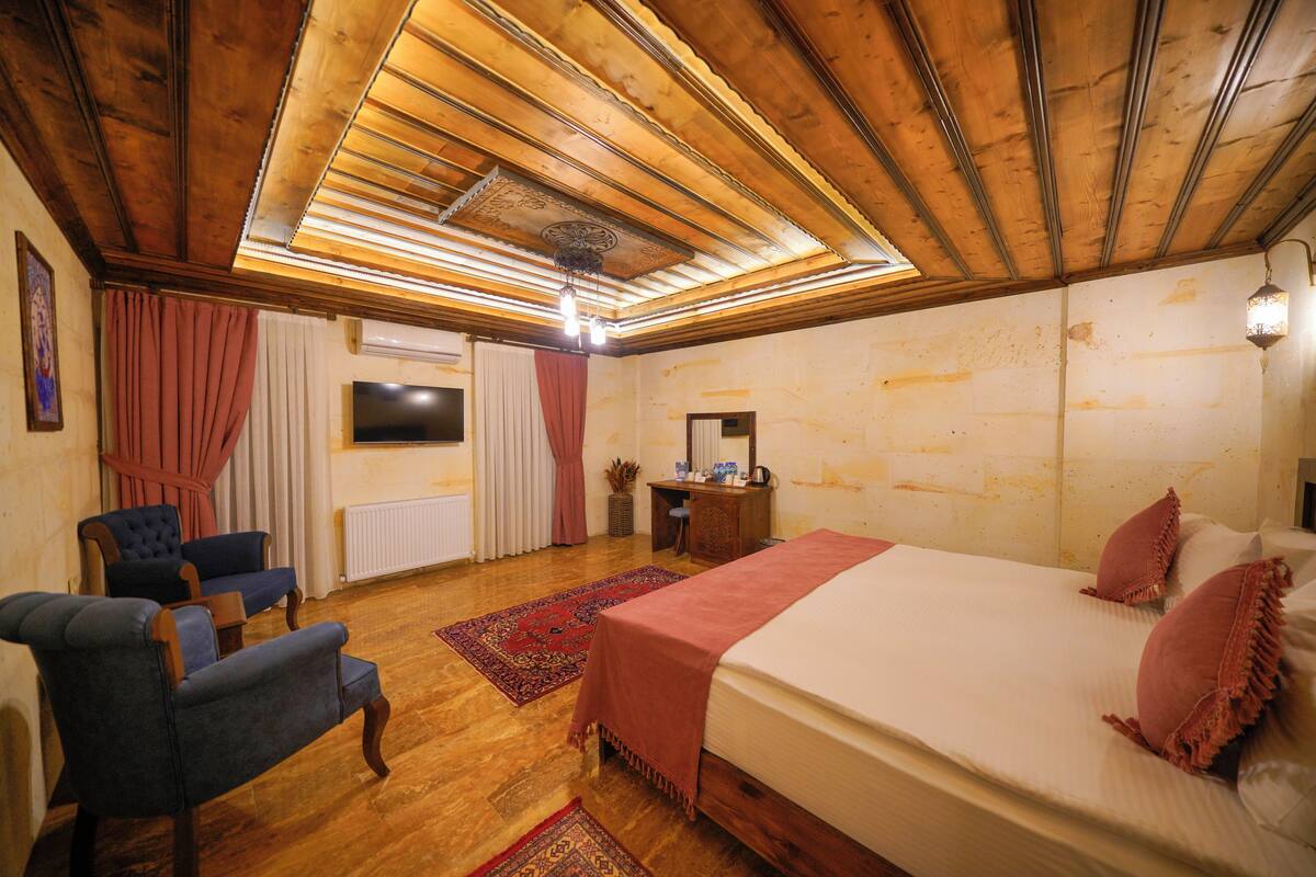 Un dormitorio con techo de madera, una cama con manta rosa, dos sillones azules, una alfombra roja y un aparador con espejo.