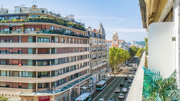 Un immeuble à plusieurs étages comprenant des logements et des espaces commerciaux, une rue avec des voitures et un autobus, et une tour de l’horloge imposante en arrière-plan.