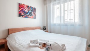 Un lit bien fait, avec du linge de lit blanc, un tableau abstrait aux couleurs vives accroché au mur et une fenêtre ornée de rideaux légers.