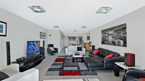 Een moderne woonkamer met een flatscreen televisie, een zwarte leren bank, een witte fauteuil en een eethoek op de achtergrond.