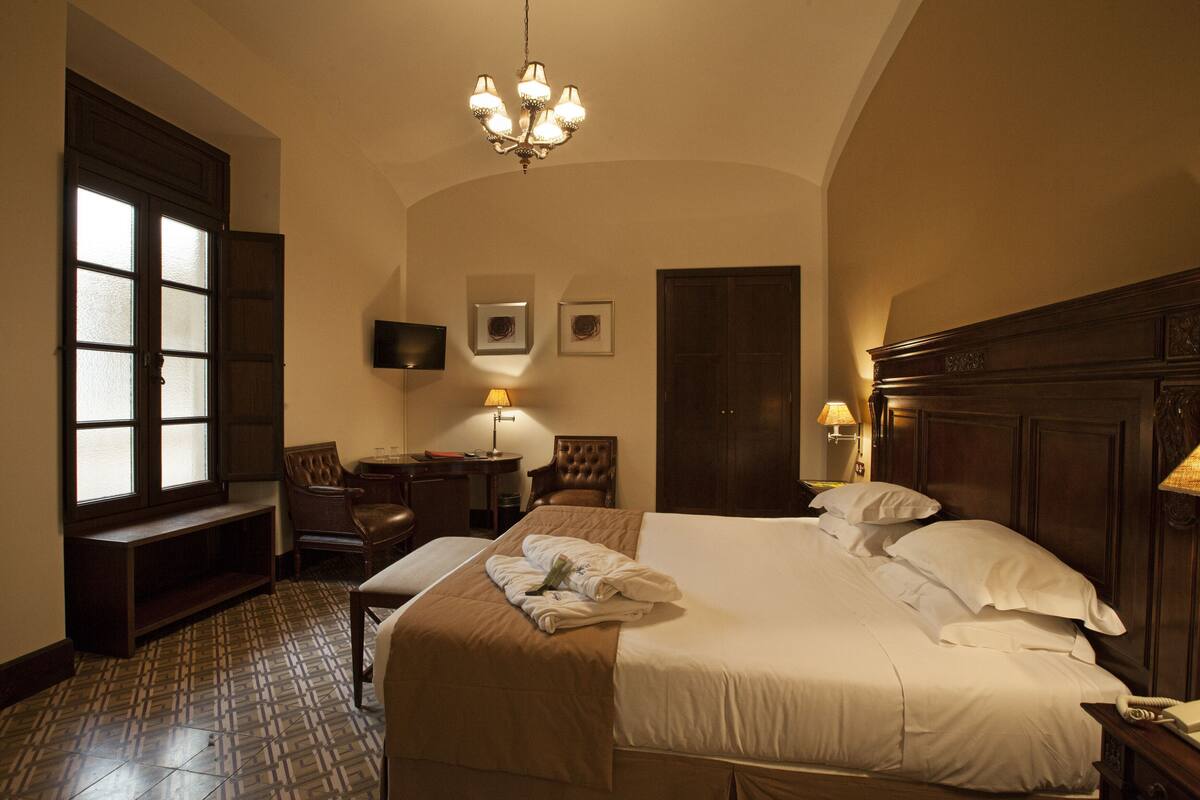 A hotel room with a large bed, a desk, a chair, a television, and a chandelier.