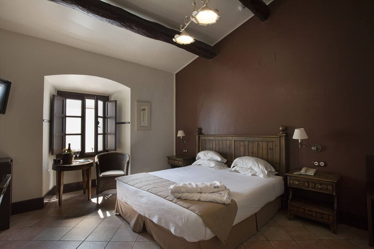 A hotel room with a large bed, a chair, a small table with a bottle and glasses, a lamp, and a telephone on a nightstand.