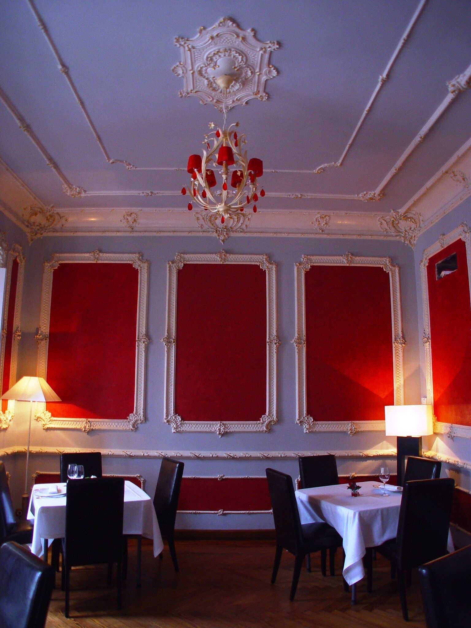 Un espace repas comprenant trois tables, chacune recouverte d’une nappe blanche et entourée de chaises noires. Les murs sont ornés de panneaux rouges et de motifs dorés travaillés.