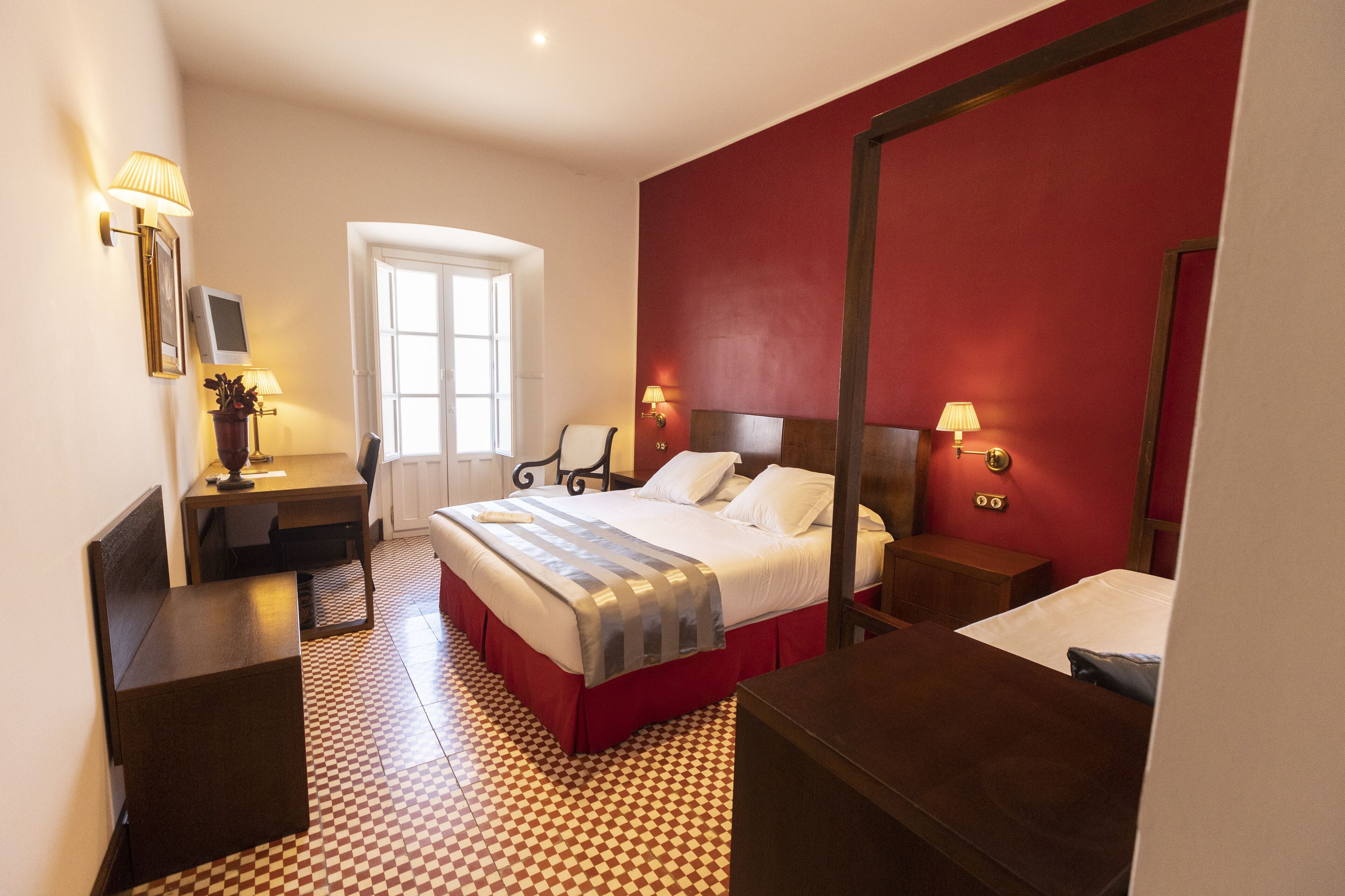 Een hotelkamer met een zwart-wit betegelde vloer, een bed met een rode hoofdbord, een bureau met een lamp en een raam met witte gordijnen.