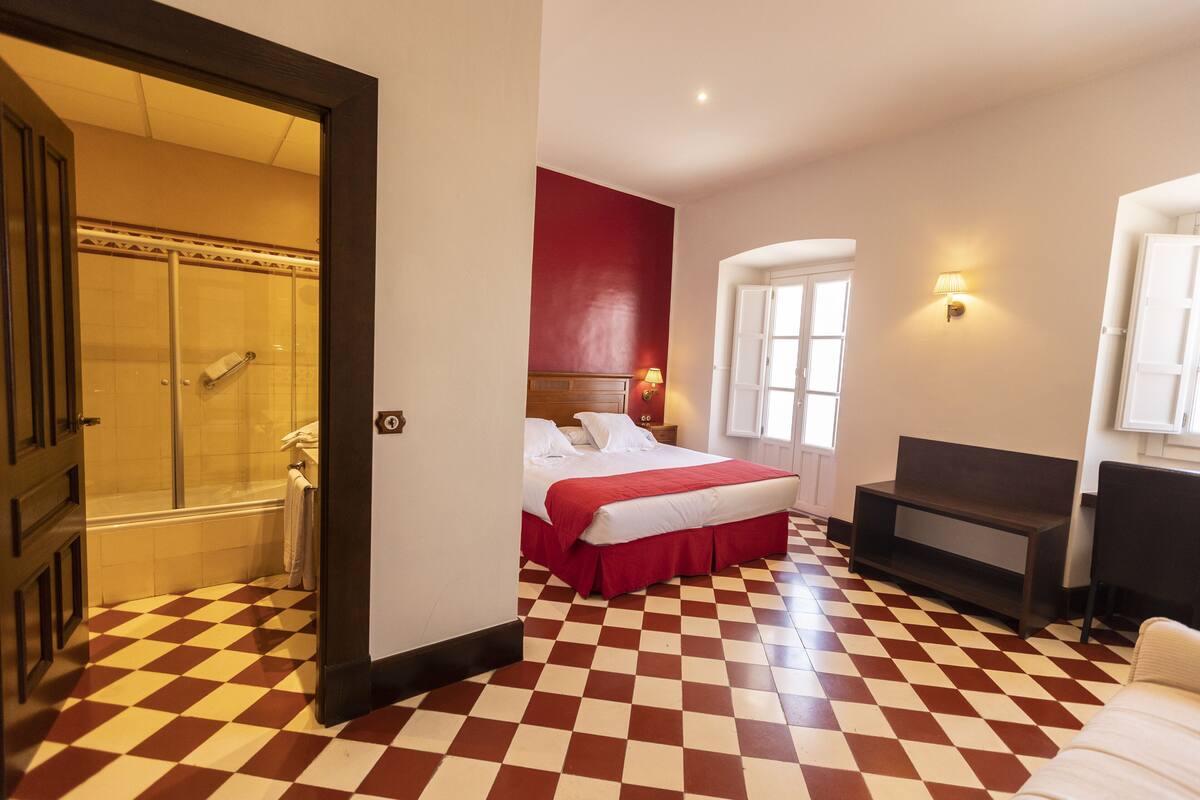 A hotel room with a checkered floor, a bed with a red headboard, a seating area, and a shower.