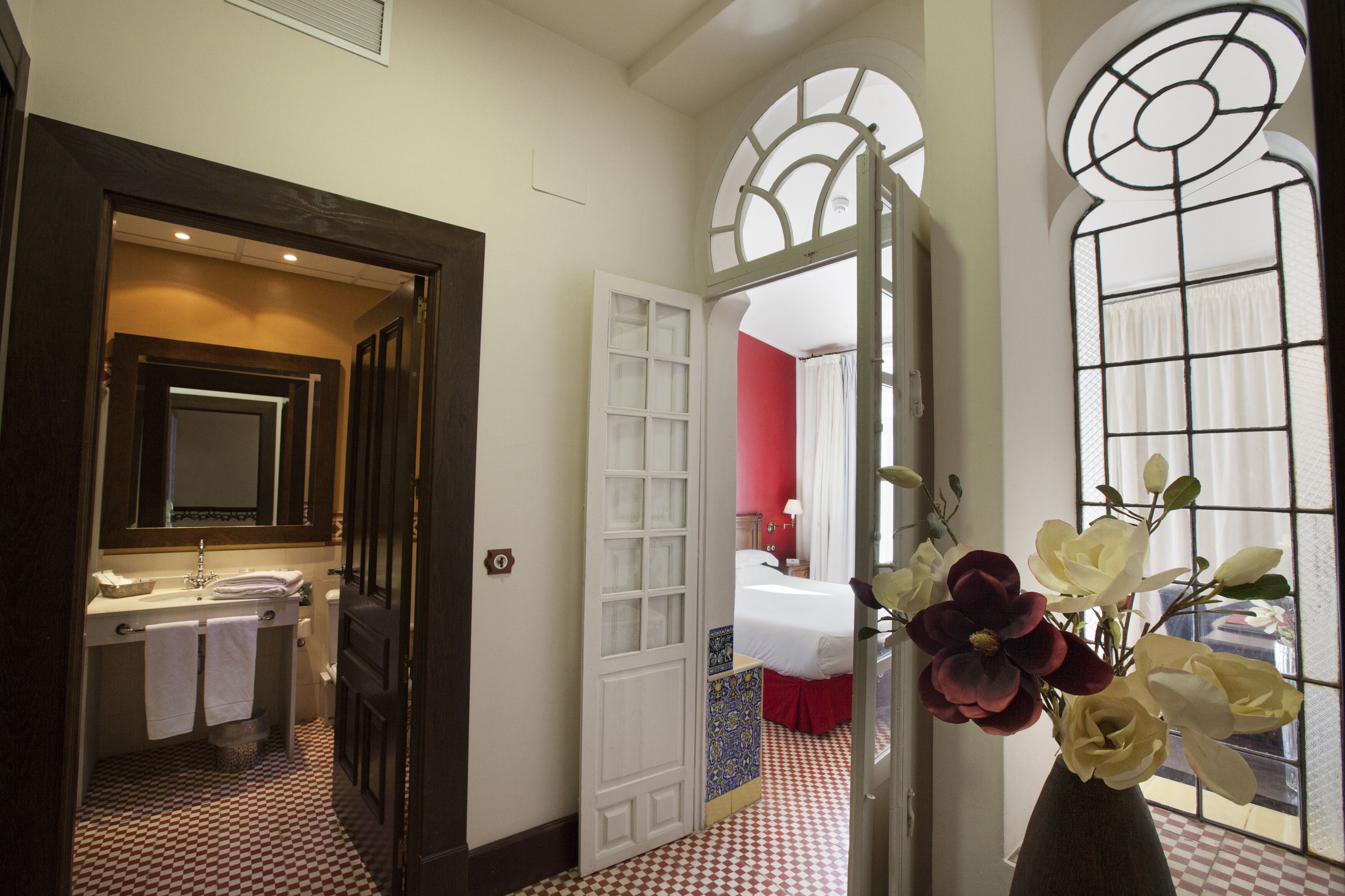 Habitación de hotel con piso a cuadros, un espejo grande, tocador con lavamanos y un jarrón con flores.