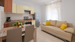 A compact kitchen with a dining area, a sofa with yellow cushions, and a window with sheer curtains. - Apartments Ena (Novalja)