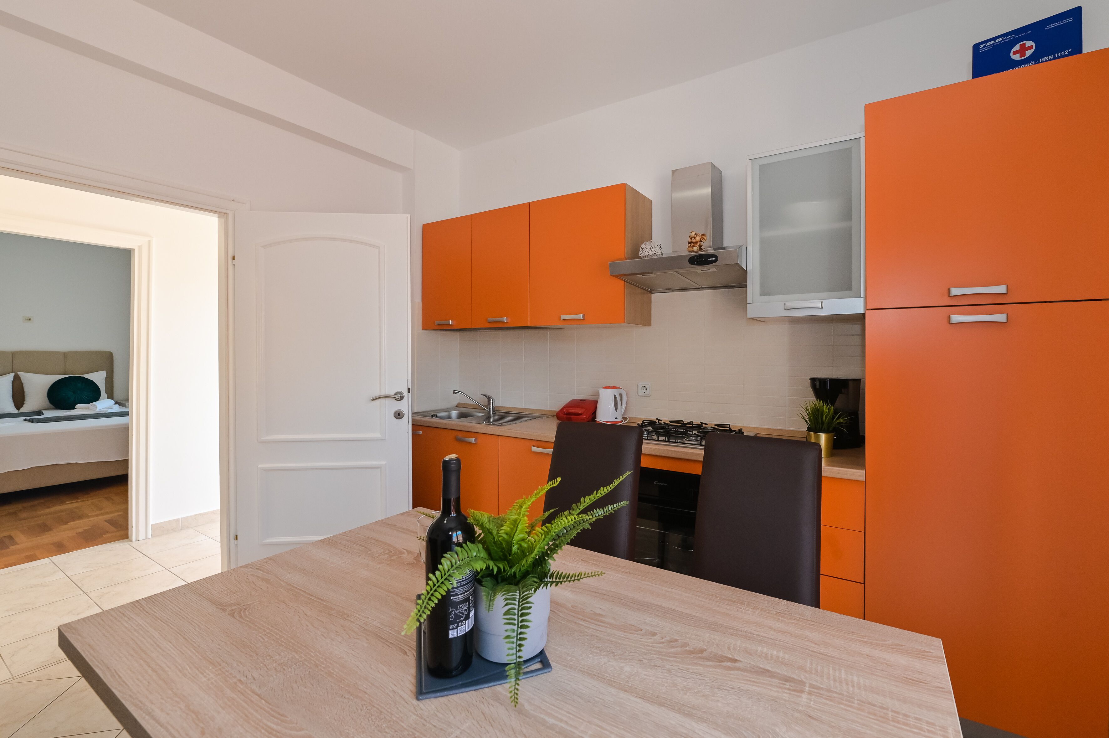 A modern kitchen with orange cabinets, a white refrigerator, and a dining table with a bottle and plant.