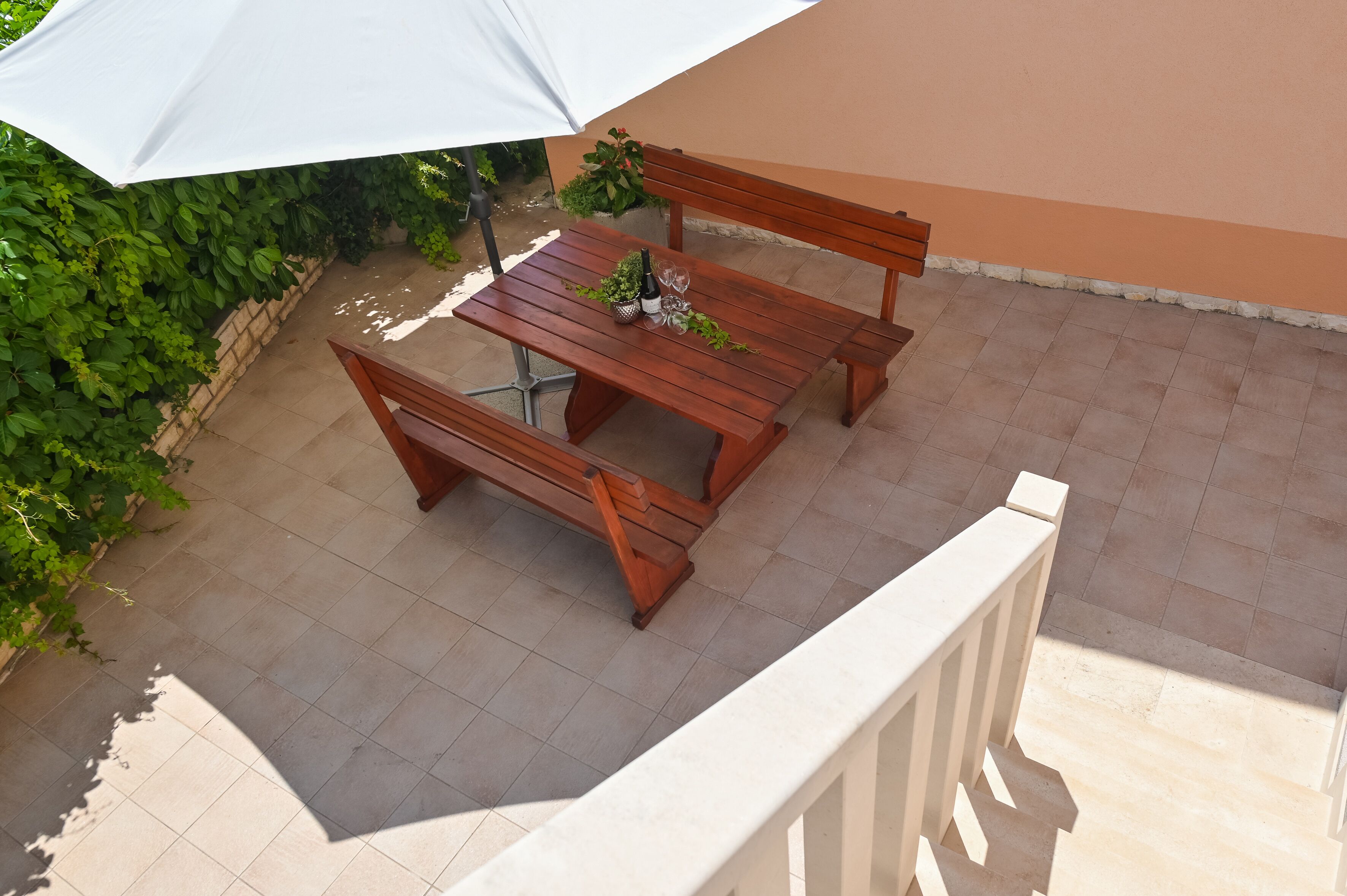 Ein Holz-Tisch-und-Bank-Set auf einer gefliesten Terrasse, mit einem weißen Sonnenschirm.