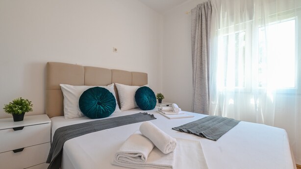A neatly made bed with white and teal pillows, a folded white towel, and a gray blanket on a white mattress in a well-lit room with a window and curtains. - Apartments Ena (Novalja)