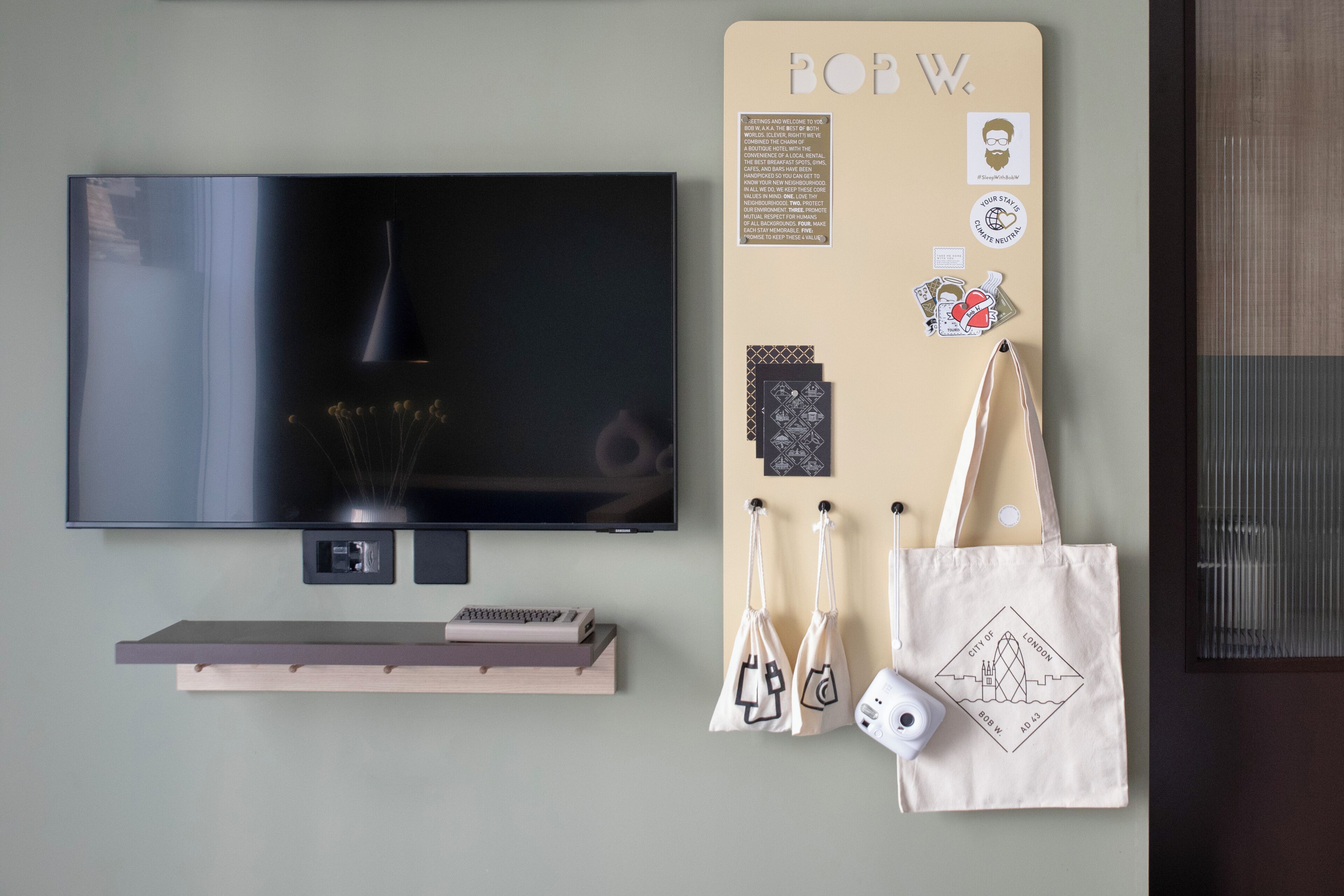 A flat-screen TV mounted on a wall, a small shelf with books and a remote control, and a bulletin board with various items including a tote bag and a camera.