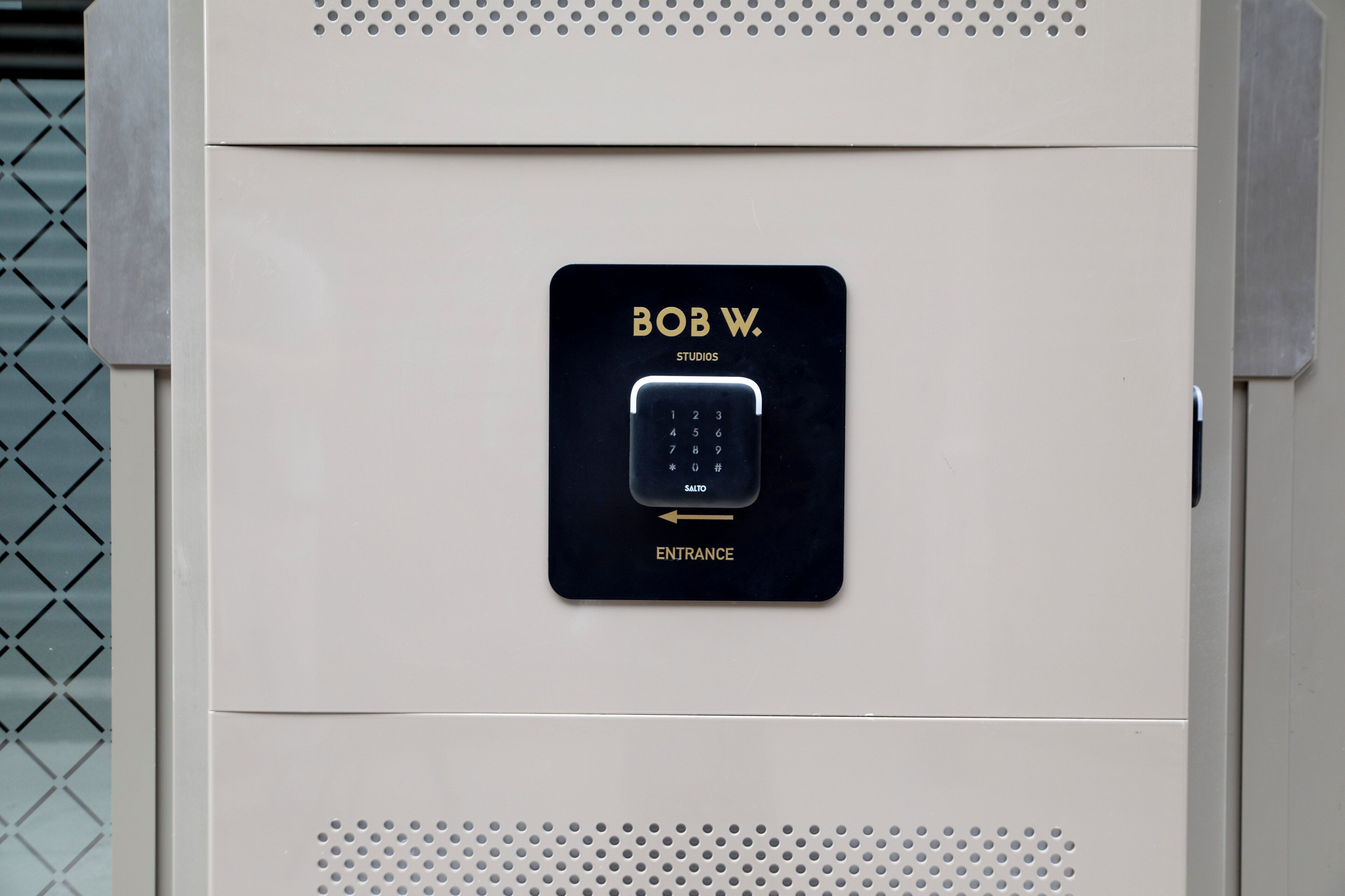 En knappsats med texten ”BOB W. STUDIOS” och ”INGÅNG”, monterad på ett beigefärgat metalls skåp med perforerade paneler.