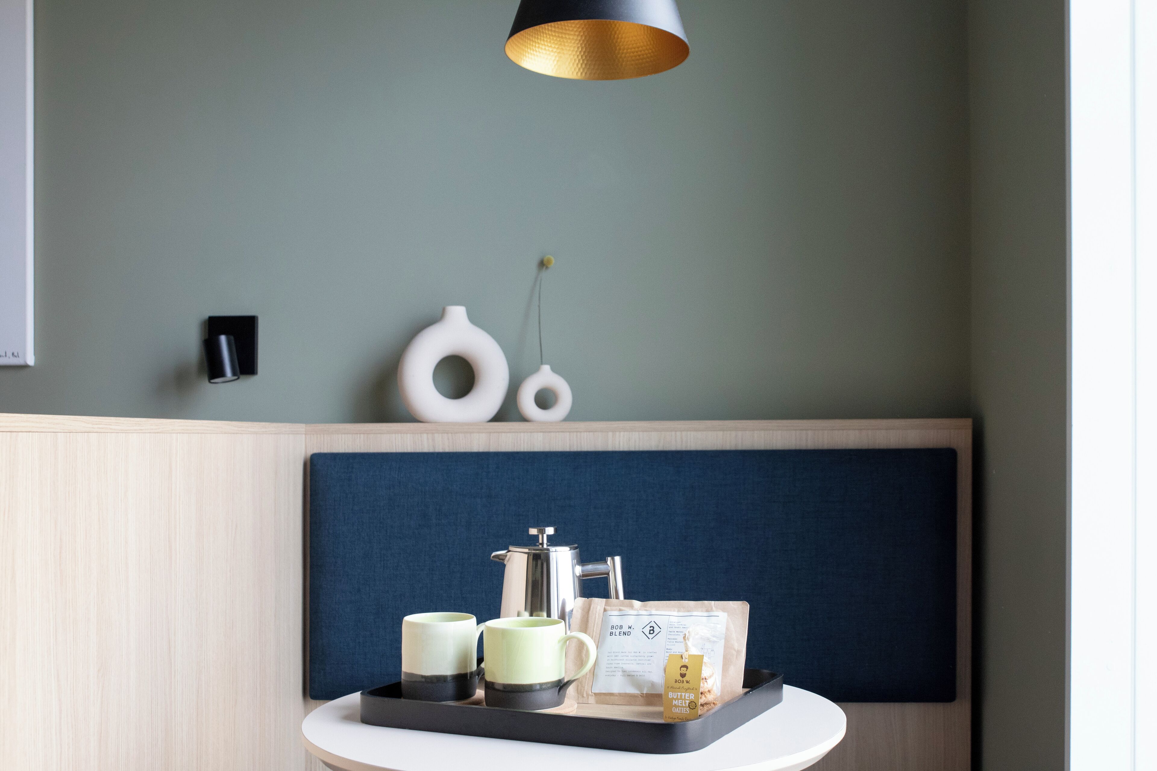 Una habitación moderna con un panel de madera en la pared, un banco acolchado azul, una mesa redonda con una bandeja que contiene una cafetera, dos tazas y una bolsita de té.