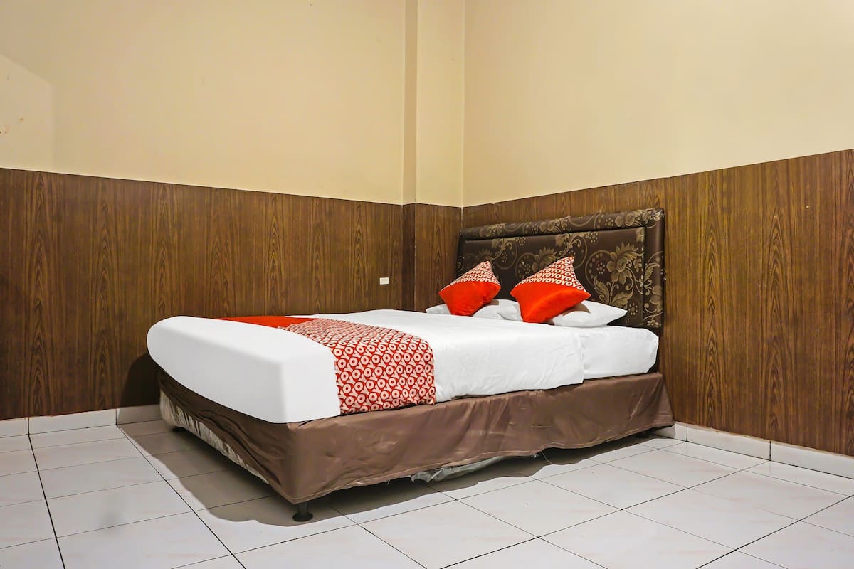 A neatly made bed with a patterned bedspread and pillows, set against a wooden wall in a corner of a room with tiled flooring.