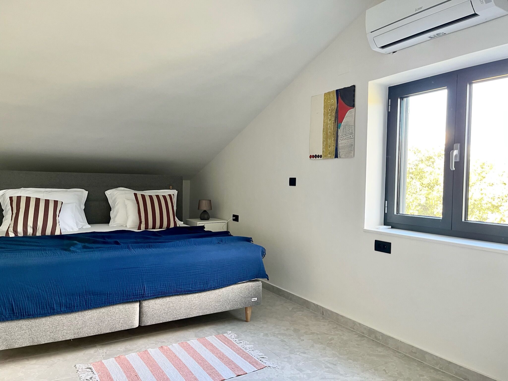 A bedroom with a bed, a window, an air conditioner, and a wall-mounted artwork.