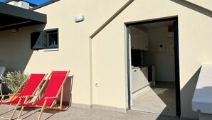 A building with a beige exterior, a small window, and a door leading to a kitchen area. There are two red lounge chairs outside. - Penthouse apartment (Šimuni)