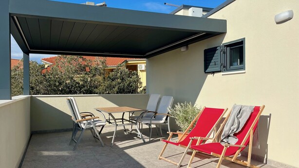 A covered outdoor seating area with a table and chairs, a potted plant, and a view of neighboring buildings. - Penthouse apartment (Šimuni)