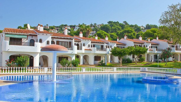 A swimming pool surrounded by white buildings with red-tiled roofs, a white fence, and greenery in the background. - Apartment SON BOU, 67 (Son Bou)