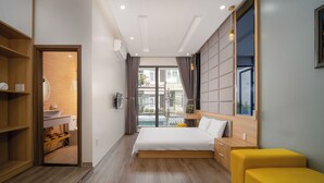 A modern hotel room with a large bed, a wooden wardrobe, a yellow ottoman, and a view of the city through the window. - N to M House (Da Nang)