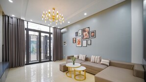 A modern living room with a beige sofa, two gold-framed coffee tables, a chandelier, and a large window with curtains. - N to M House (Da Nang)