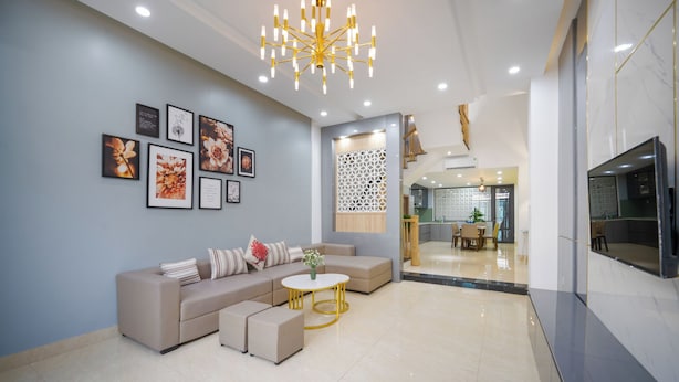 A modern living room with a beige sectional sofa, a round gold table, and a decorative wall with framed floral artwork. - N to M House (Da Nang)