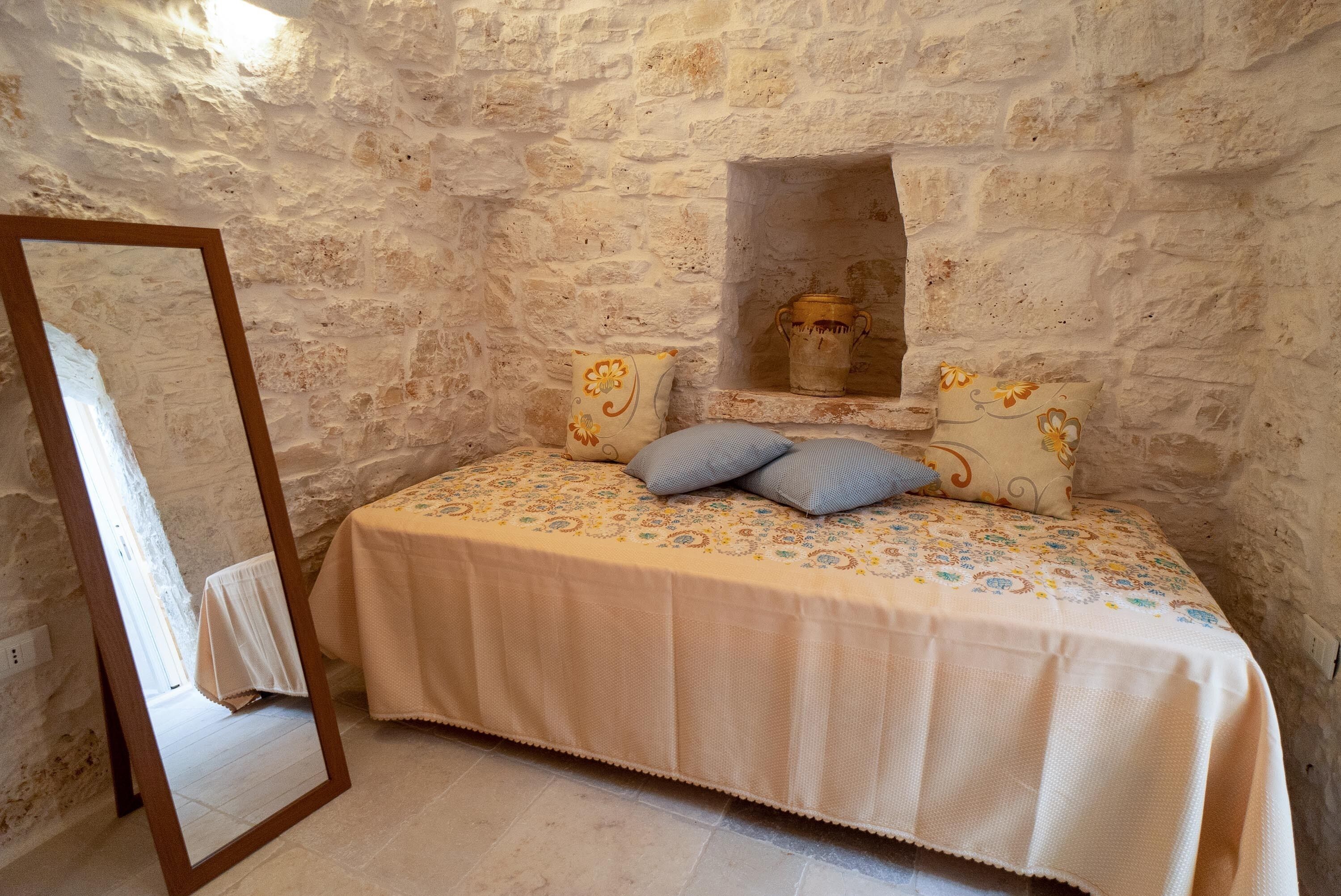 Un dormitorio con pared de piedra, cama con ropa de cama estampada, espejo y estante con objetos decorativos.