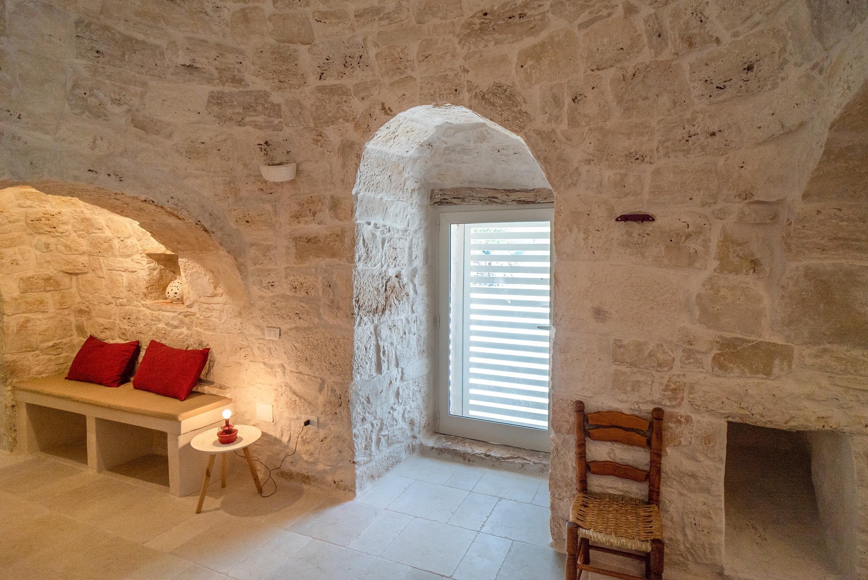 Une pièce aux murs de pierre, avec un banc, une petite table sur laquelle est posée une bougie et une chaise en bois.