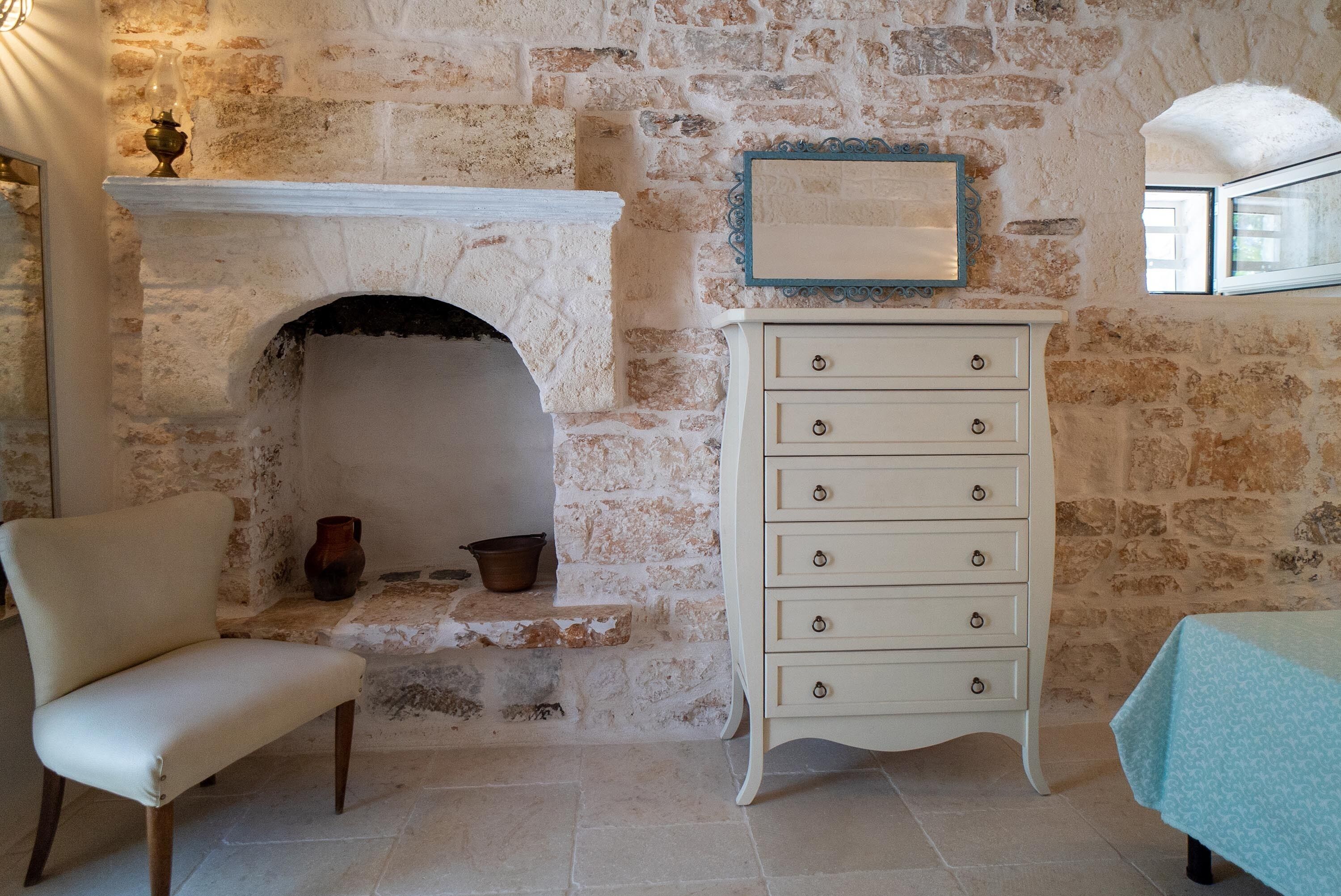 Une pièce aux murs de pierre, avec une cheminée, une commode blanche, une chaise et une table recouverte d’une nappe bleue.