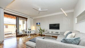 Una sala de estar moderna con un sofá gris, piso de madera, un ventilador de techo, un televisor y un comedor con sillas y una mesa.