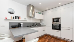 Una cocina moderna con armarios blancos, una campana extractora de acero inoxidable, microondas integrado y horno. Hay dos taburetes blancos y suelo de madera.