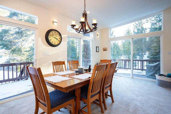 Comedor con mesa de madera, sillas, una lámpara de araña y ventanas amplias con vistas a árboles.