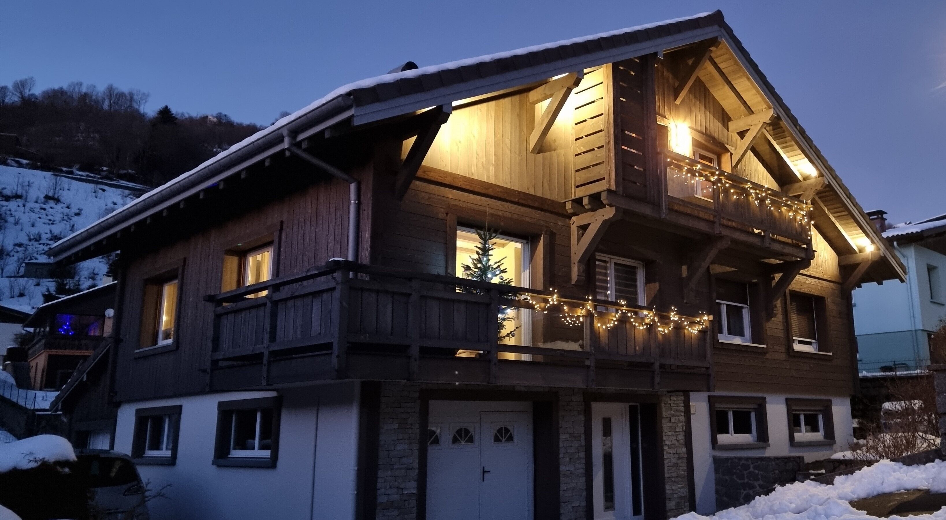 A two-story wooden chalet with a balcony, a small balcony with string lights, and a snow-covered roof.