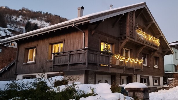 Een houten chalet met veel ramen, een balkon met lichtjes en een besneeuwde omgeving.