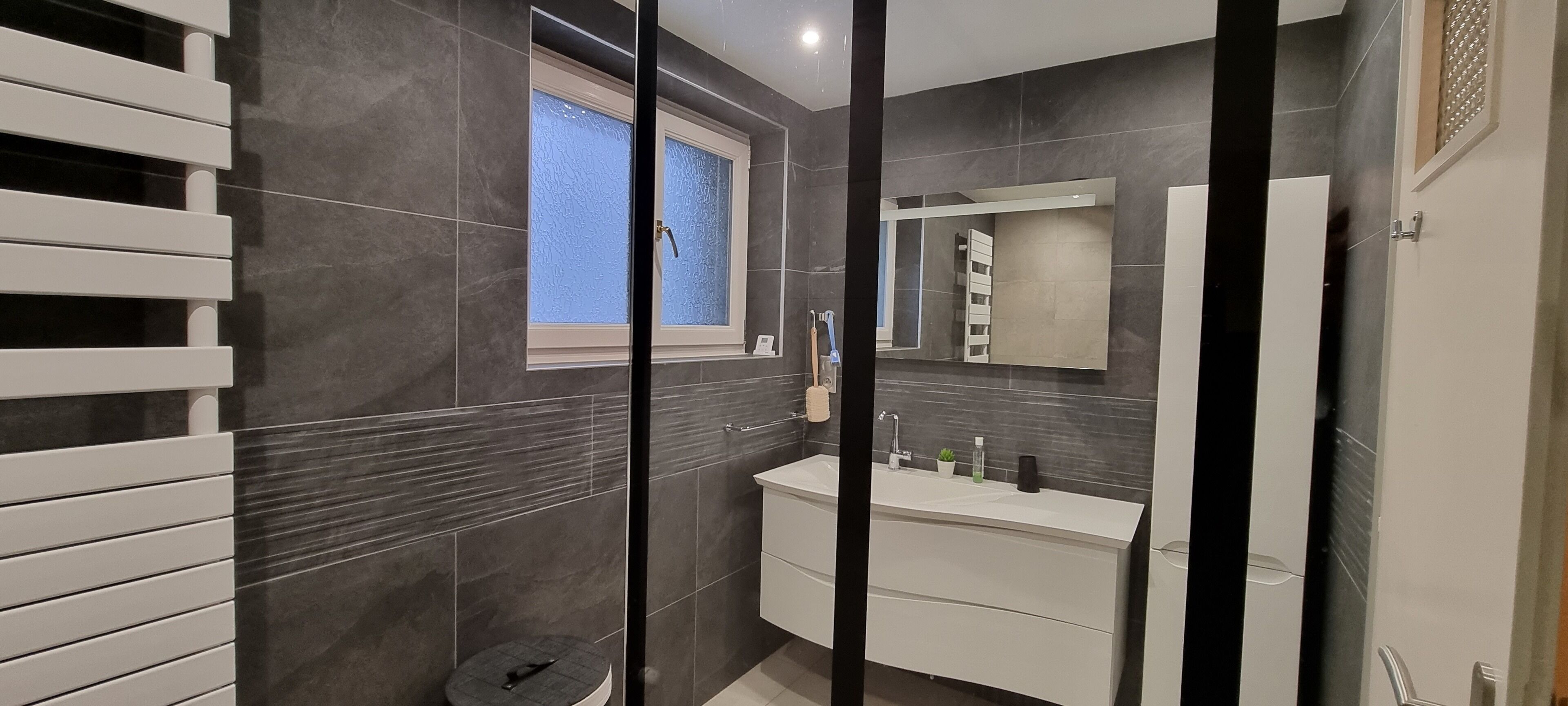 Een moderne badkamer met donkere betegelde wanden, een witte wastafel met spiegel, en een raam met gezandstraald glas.