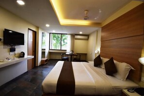 A hotel room with a bed, a TV, a desk, and a window with a view of trees. - Hotel Pearl  (Kolhapur)