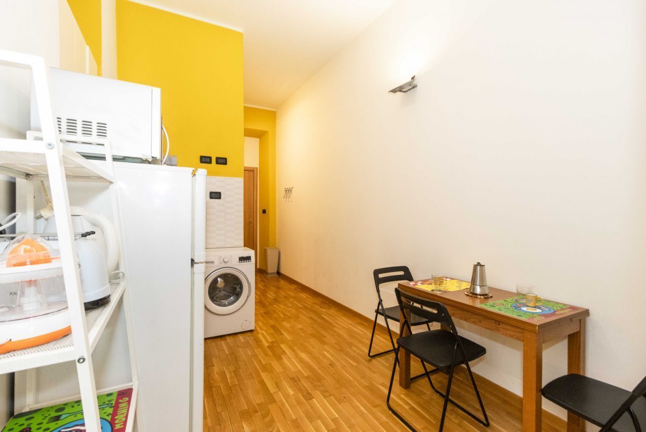 Una habitación compacta con lavadora, una mesita con sillas y un estante con diversos objetos.