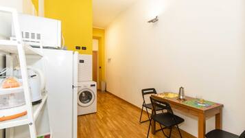Una habitación compacta con lavadora, una mesita con sillas y un estante con diversos objetos.